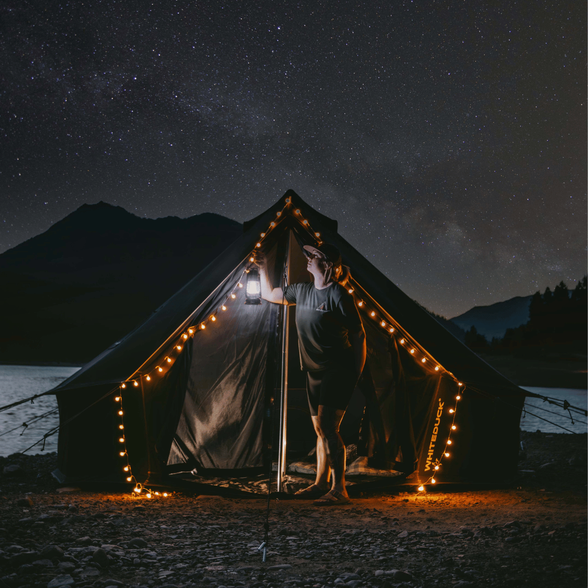 Person standing in front of a tent with string lights under a starry night sky.