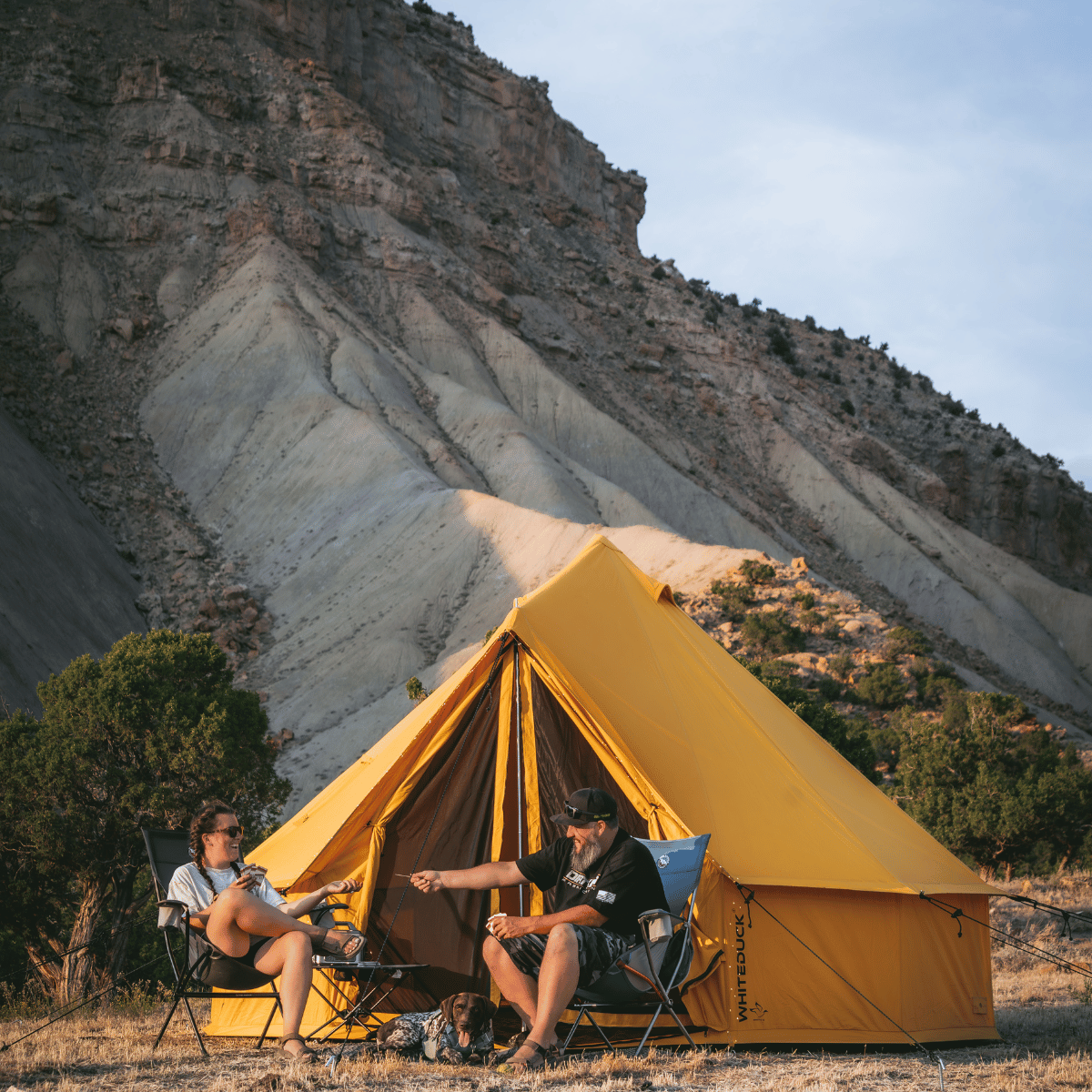 Two people sitting outside a yellow tent with a mountainous landscape in the background