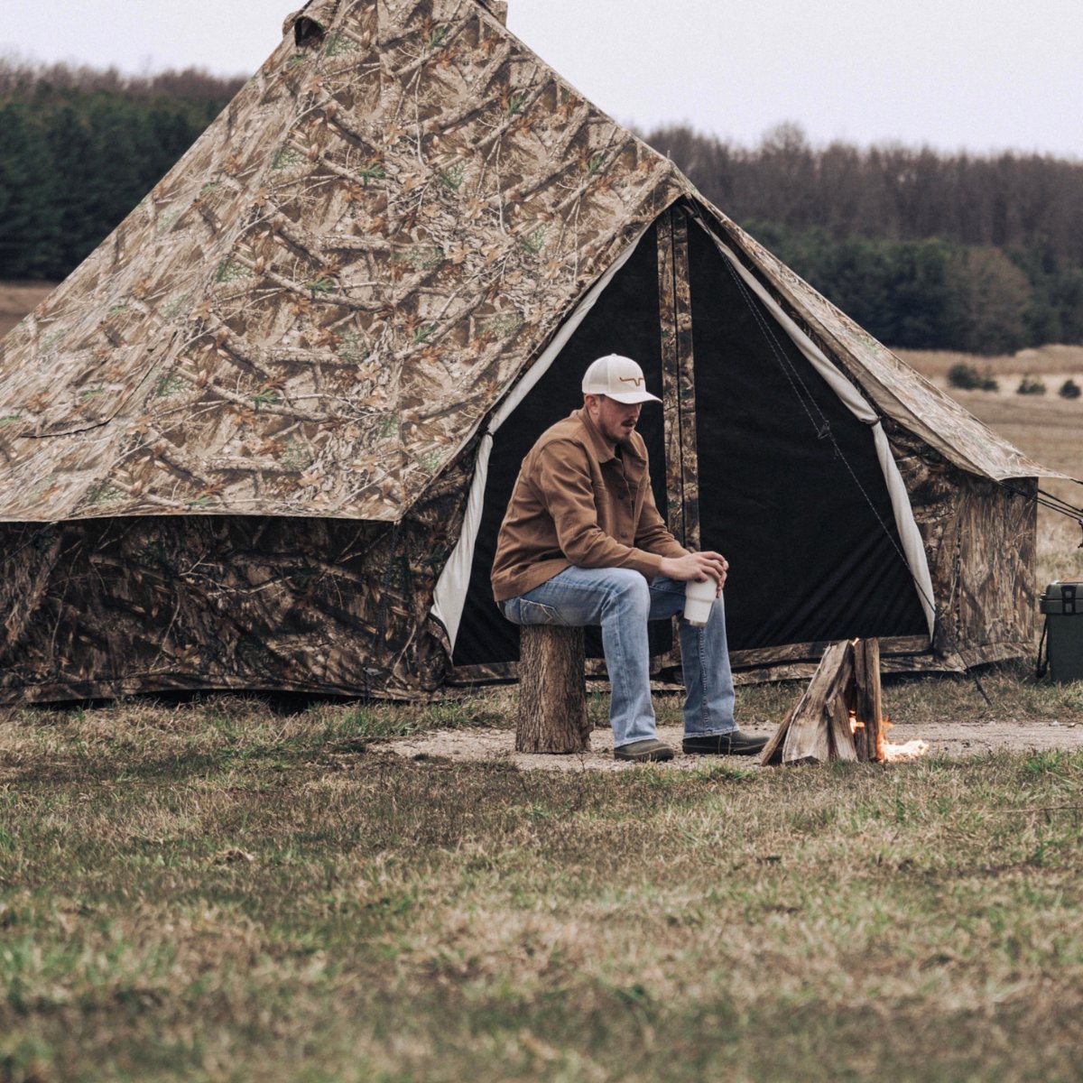 Man sitting by a campfire in front of a camouflage tent in a field.