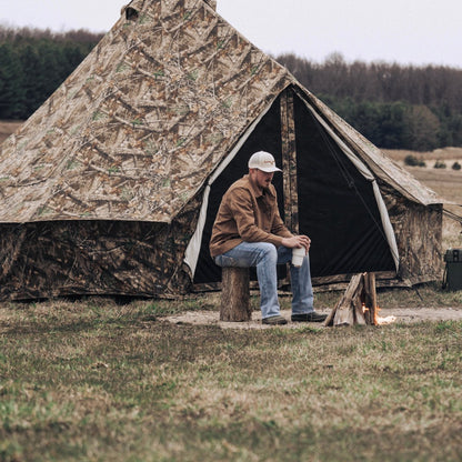 Man sitting by a campfire in front of a camouflage tent in a field.